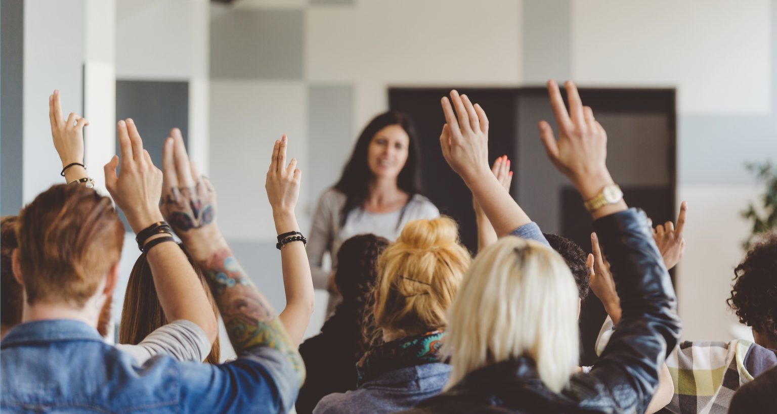 Back view of large group of students attending a seminar at the university, raising their hands and voting. Focus on hands and heads. Teacher in the background.