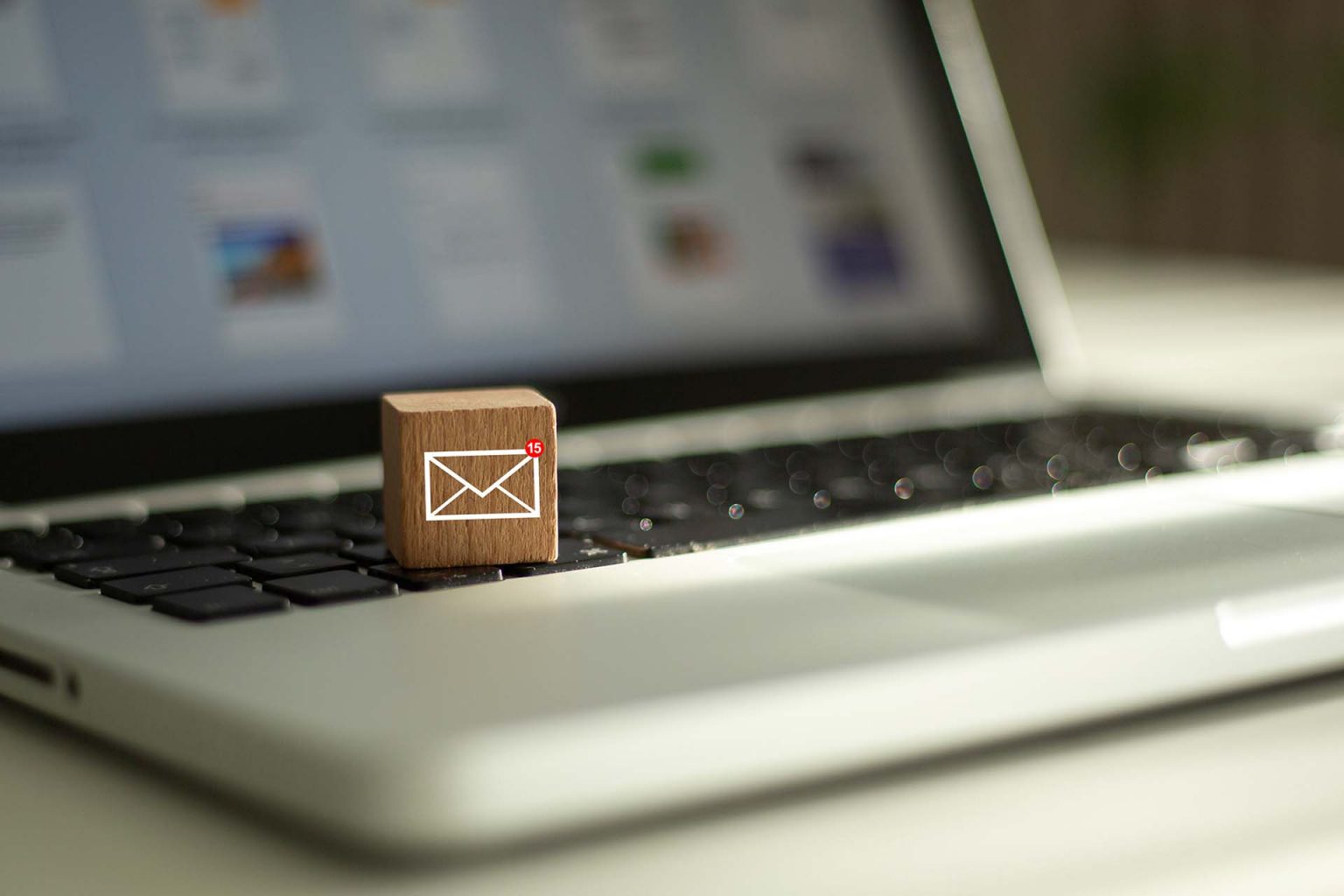 Wooden block sitting on the keyboard of a grey laptop. On the front of the wooden block is an icon of an envelope with the number 15 in a small red circle on the top left. This icon represents a new email.