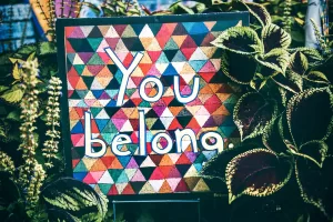 A colourful painted sign that says you belong surrounded by plants.
